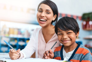 Portrait, teacher and child in class together for help in growth, development and support for writing in education. Learning, woman and boy at desk with notes, smile and lesson at elementary school Portrait, teacher and child in class together for help in growth, development and support for writing in education. Learning, woman and boy at desk with notes, smile and lesson at elementary school.